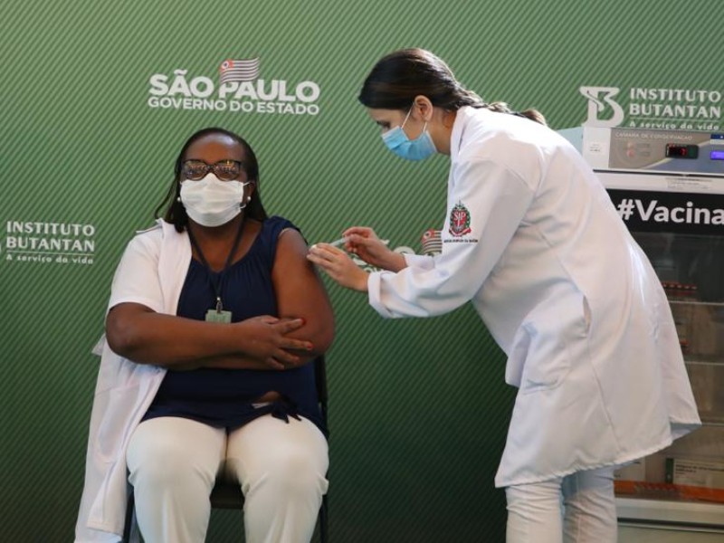 Mônica está sentada enquanto uma enfermeira aplica a primeira dose da vacina contra a covid-19 em seu braço esquerdo. No canto direito aparece uma geladeira onde estão armazenadas as vacinas